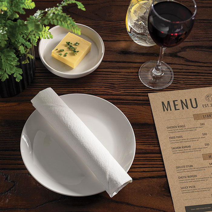 White linen-like napkin with rolled cutlery inside displayed on a plate and wooden table surrounded by a glass of wine, butter and menu