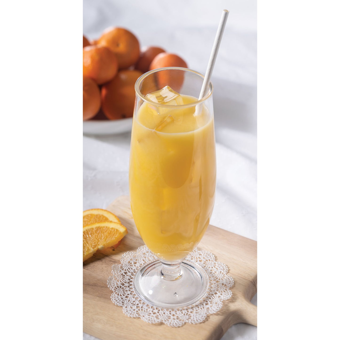 Glass of orange juice with a white straw, surrounded by oranges on a wood cutting board
