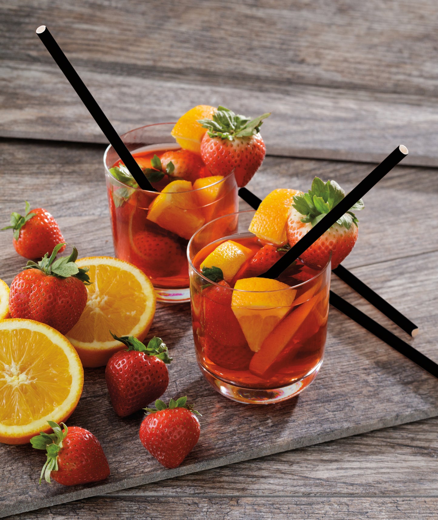 Two glasses of red fruit punch with strawberries and oranges and black paper straws on a wooden surface
