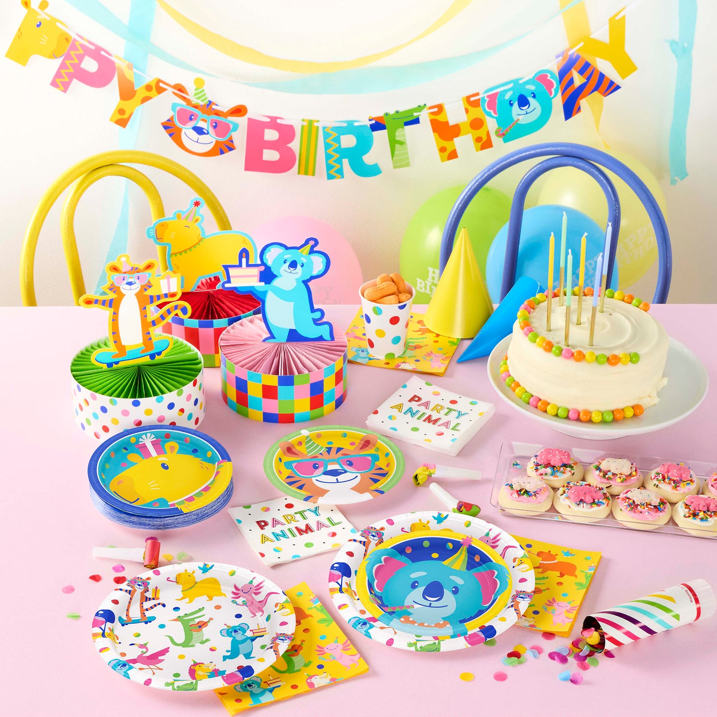Birthday party setup with colorful decorations, cake, and plates on a pink tablecloth.