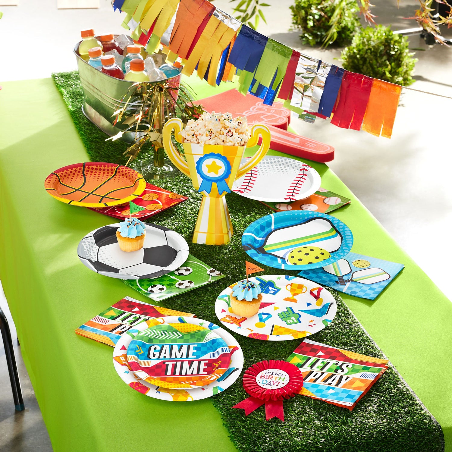Sport themed party supplies displayed on a green table with basketball, baseball, soccer, and pickleball-themed plates and napkins
