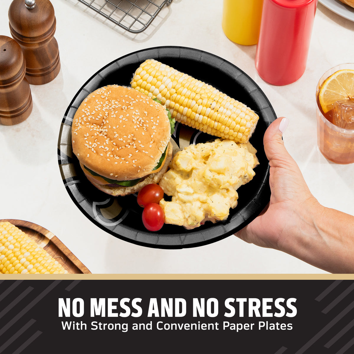 A hand holds a black New Orleans Saints paper plate with a hamburger, corn on the cob, potatoes, and cherry tomatoes on a kitchen counter. Condiment bottles, pepper mills, and a drink are visible in the background, with the text 'NO MESS AND NO STRESS With Strong and Convenient Paper Plates' at the bottom.
