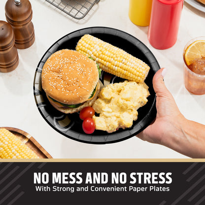 A hand holds a black New Orleans Saints paper plate with a hamburger, corn on the cob, potatoes, and cherry tomatoes on a kitchen counter. Condiment bottles, pepper mills, and a drink are visible in the background, with the text 'NO MESS AND NO STRESS With Strong and Convenient Paper Plates' at the bottom.