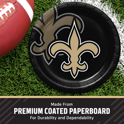 A black paper plate featuring the New Orleans Saints fleur-de-lis logo, shown on a grassy field with a football partially visible. Text at the bottom reads 'Made From PREMIUM COATED PAPERBOARD For Durability and Dependability'.