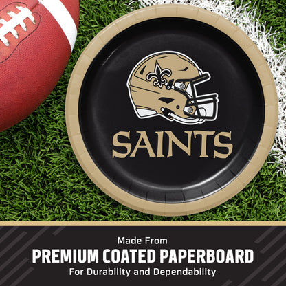 A round black paper plate with a gold rim, printed with the New Orleans Saints helmet logo and the word 'SAINTS' in gold, shown on a grassy field with a football partially visible. Text at the bottom reads 'Made From PREMIUM COATED PAPERBOARD For Durability and Dependability'.