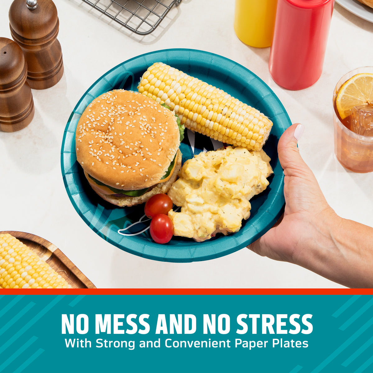 A teal paper plate being held by a hand, filled with a hamburger, corn on the cob, potato salad, and cherry tomatoes, set on a kitchen counter with condiment bottles and food props. A text banner at the bottom reads 'NO MESS AND NO STRESS With Strong and Convenient Paper Plates'.