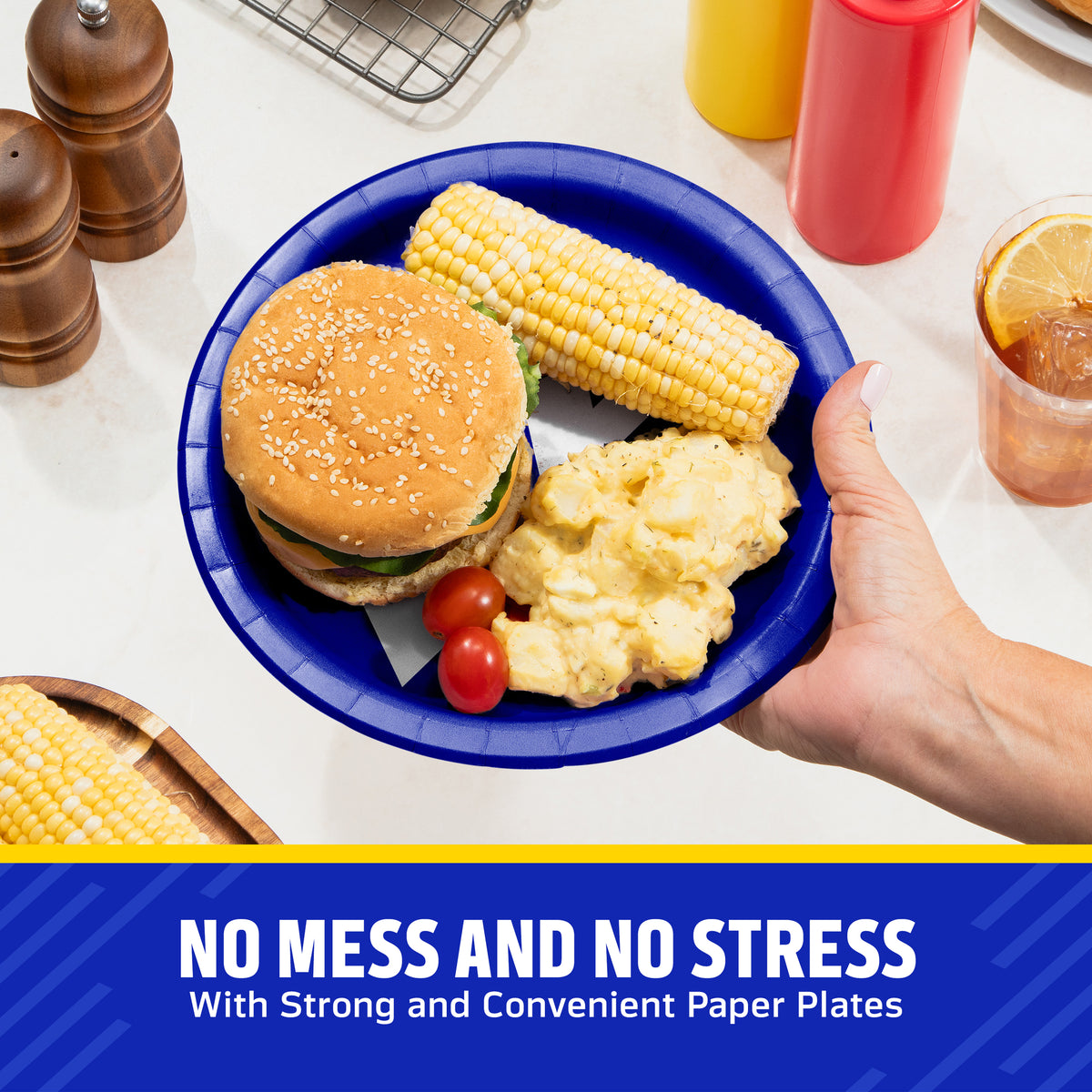 A hand holds a blue Los Angeles Rams paper plate with a hamburger, corn on the cob, mashed potatoes, and cherry tomatoes on a kitchen counter. Condiment bottles, pepper mills, and a drink are visible in the background, with a blue banner at the bottom reading 'NO MESS AND NO STRESS With Strong and Convenient Paper Plates'.