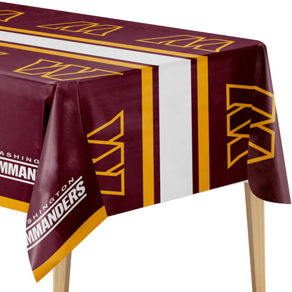 A maroon red and yellow Washington Commanders tablecloth draped over a table, featuring the team's logo and name in white text, shown against a plain white background.
