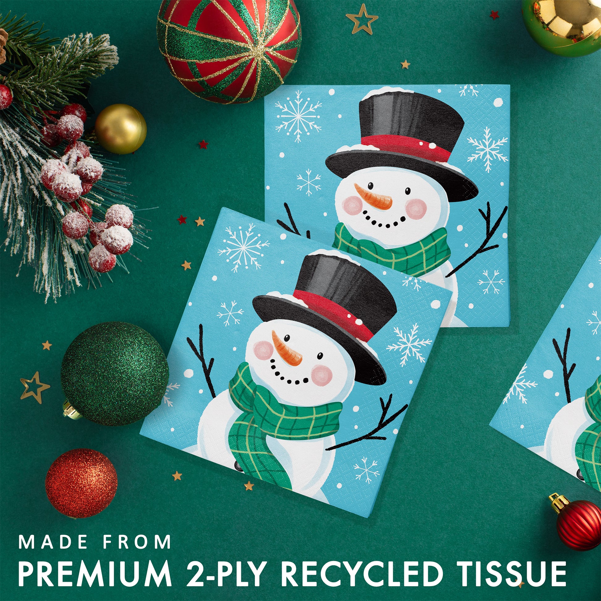 Two Christmas-themed paper napkins with a snowman and snowflakes on a blue background placed on a green background and Christmas decorations.