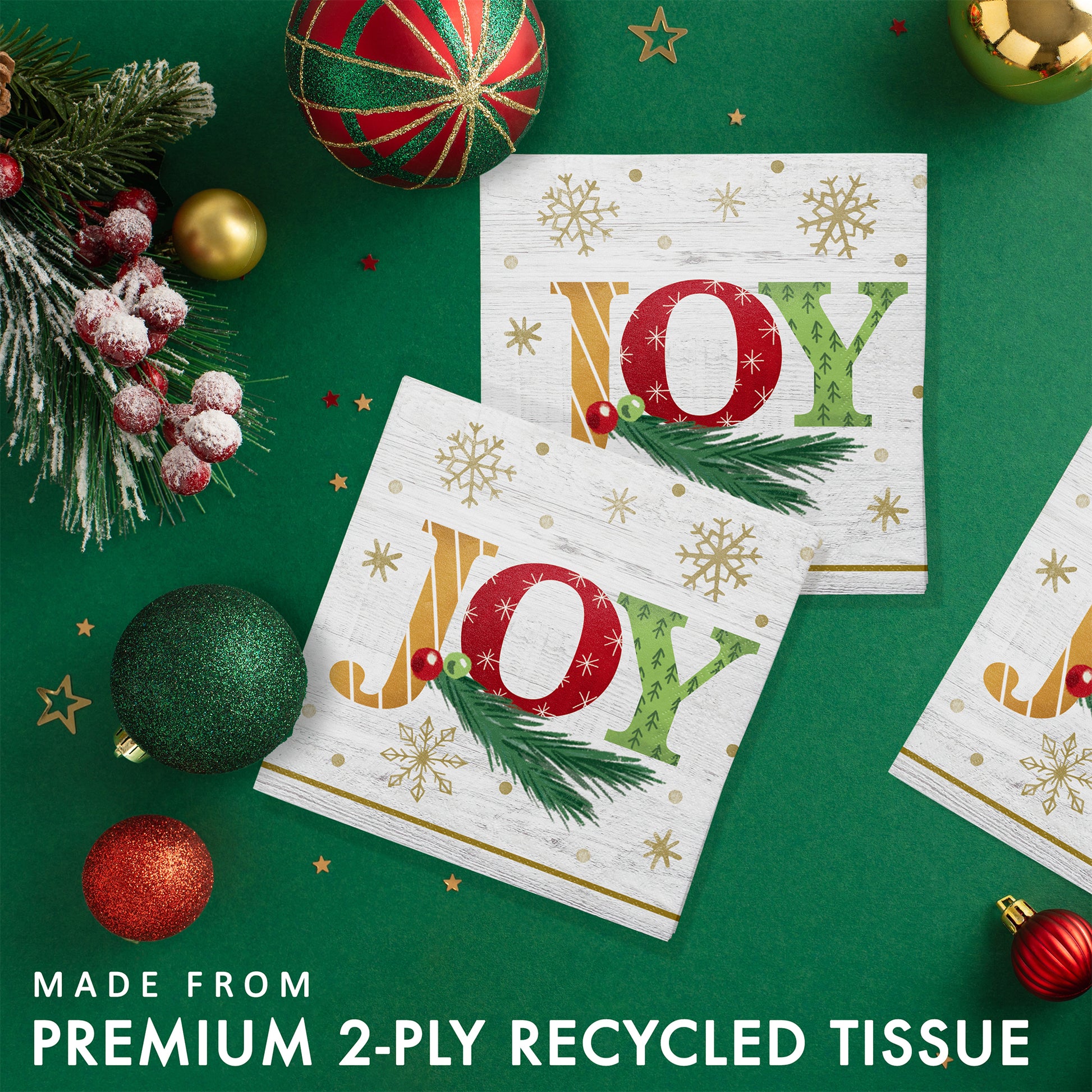 Two 'JOY' paper napkins with gold snowflakes and greenery. Napkins are shown with Christmas decorations on a green background