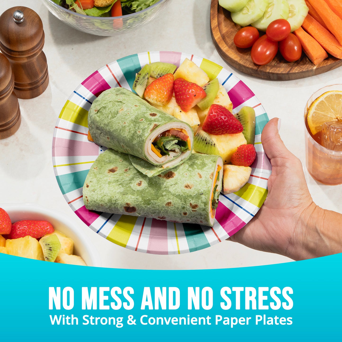 Colorful paper plate with a wrap and fruit salad, held by a hand, on a table with additional food items.