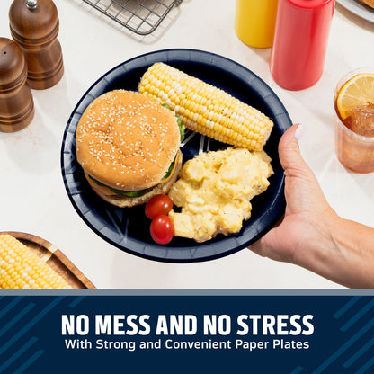 A hand holds a dark blue paper plate with a hamburger, corn cob, mashed potatoes, and cherry tomatoes on a table. Condiment bottles, pepper mills, and a drink are visible in the background, with a text banner at the bottom reading 'NO MESS AND NO STRESS With Strong and Convenient Paper Plates'.