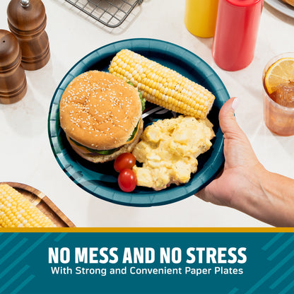 A hand holds a dark teal paper plate containing a hamburger, corn on the cob, mashed potatoes, and cherry tomatoes on a kitchen counter. Condiment bottles, pepper mills, and a drink are visible in the background, with a text banner at the bottom reading 'NO MESS AND NO STRESS With Strong and Convenient Paper Plates'.