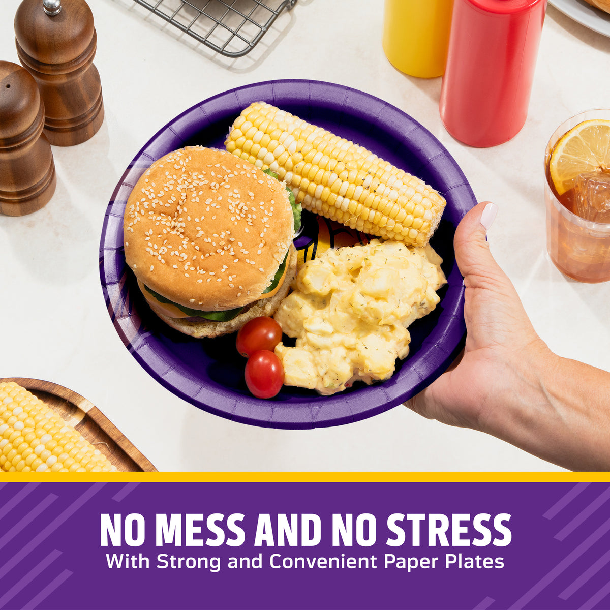 A hand holds a purple paper plate with a hamburger, corn on the cob, potatoes, and cherry tomatoes on a kitchen counter. Condiment bottles, pepper mills, and a drink are visible in the background, with a text banner at the bottom reading 'NO MESS AND NO STRESS With Strong and Convenient Paper Plates'.