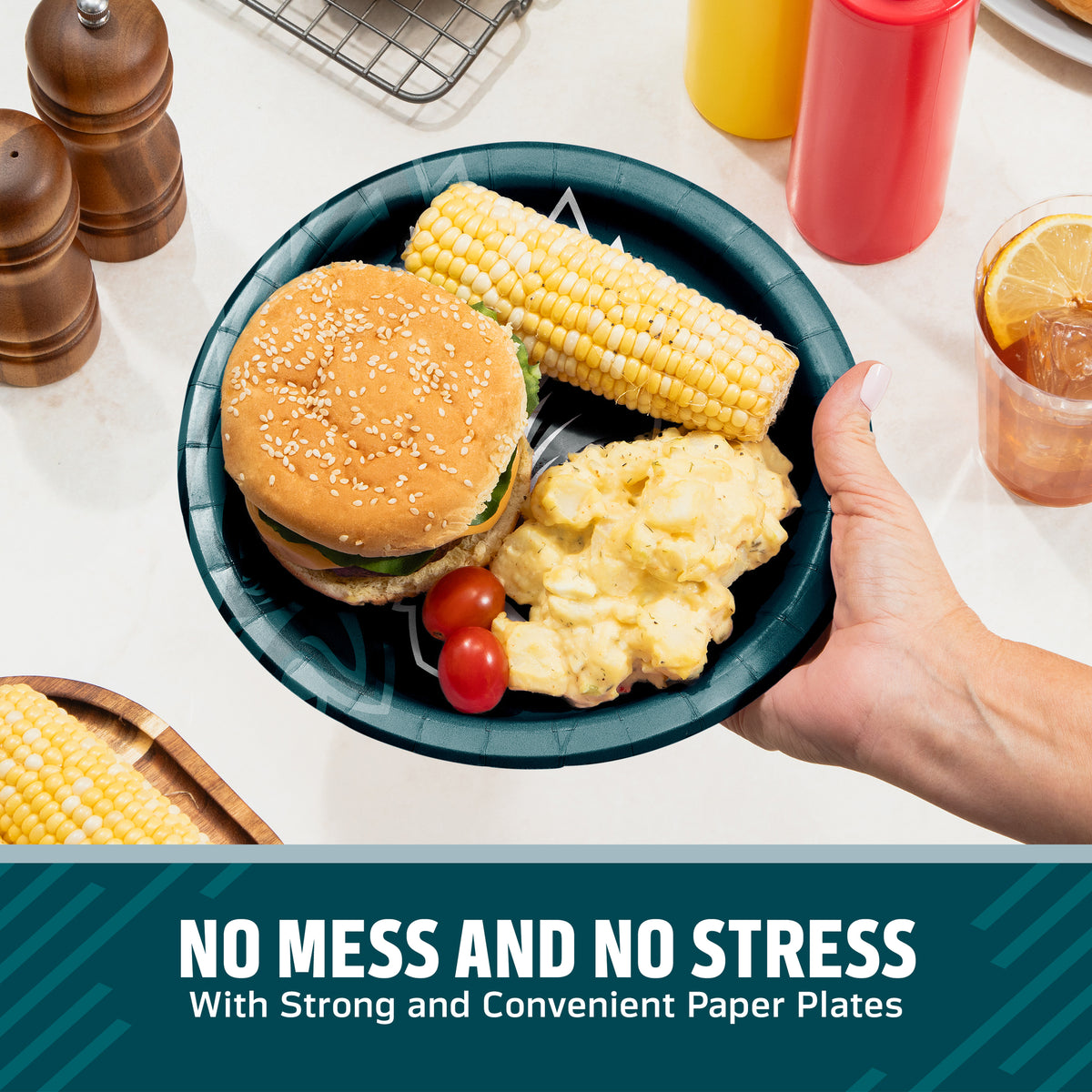 A hand holds a dark teal paper plate with a hamburger, corn on the cob, potatoes, and cherry tomatoes on a white countertop. Condiment bottles, pepper mills, and a drink are visible in the background, with a text banner at the bottom reading 'NO MESS AND NO STRESS With Strong and Convenient Paper Plates'.