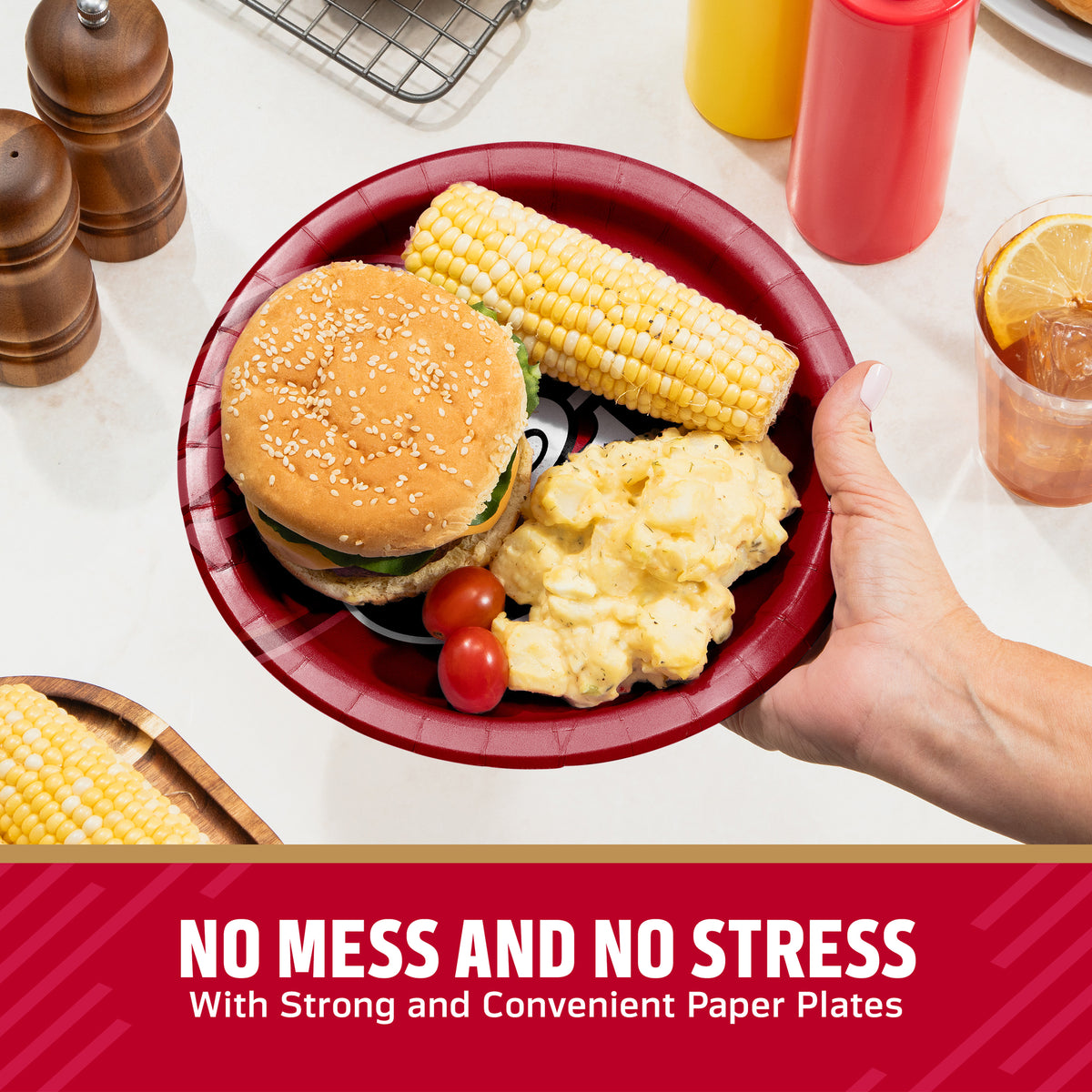 A hand holds a red paper plate with a hamburger, corn on the cob, potatoes, and cherry tomatoes on a kitchen counter. Condiment bottles, pepper mills, and a drink are visible in the background, with a text banner at the bottom reading 'NO MESS AND NO STRESS With Strong and Convenient Paper Plates'.
