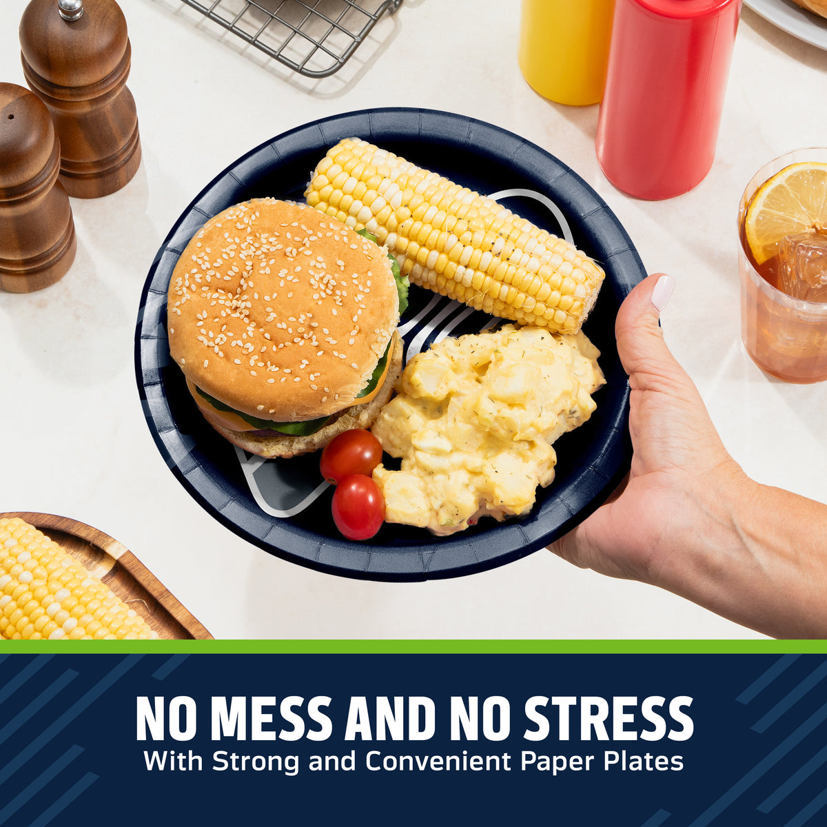 A hand holds a dark navy Seattle Seahawks paper plate with a hamburger, corn on the cob, potatoes, and cherry tomatoes on a white kitchen counter. Condiment bottles, pepper mills, and a drink are visible in the background, with a text banner at the bottom reading 'NO MESS AND NO STRESS With Strong and Convenient Paper Plates'.