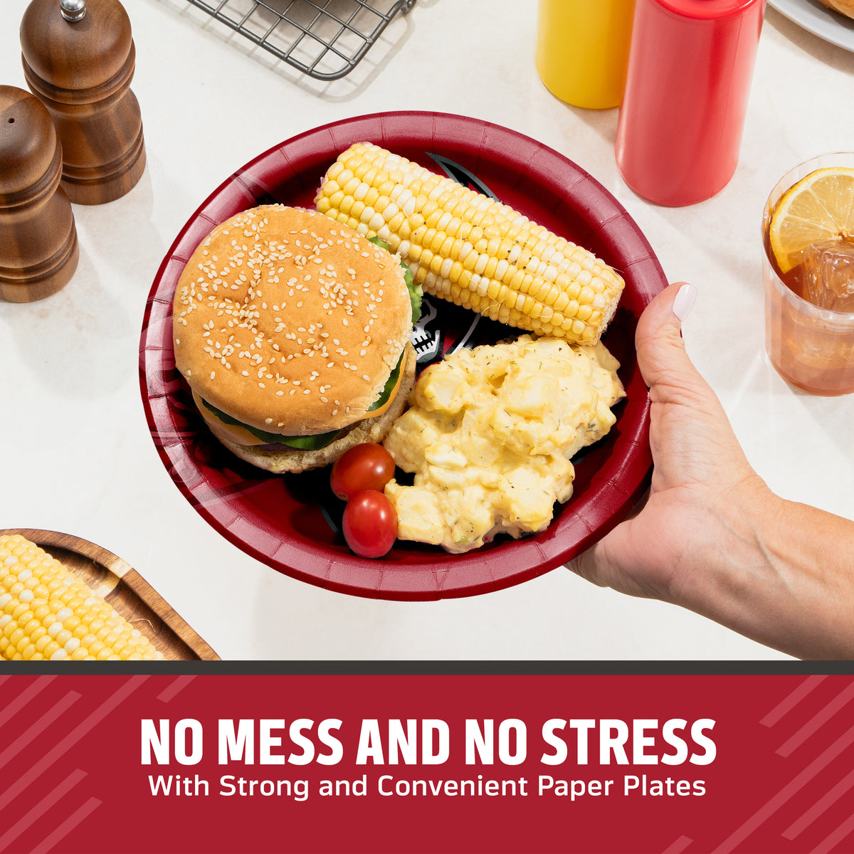 A hand holds a red paper plate with a hamburger, corn on the cob, potatoes, and cherry tomatoes on a kitchen counter. Condiment bottles, pepper mills, and a drink are visible in the background, with a banner at the bottom reading 'NO MESS AND NO STRESS With Strong and Convenient Paper Plates'.