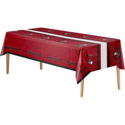 A rectangular table covered with a red tablecloth featuring repeated black, red, and white Buccaneers logos and text, plus a white vertical stripe down the center, shown on a plain white background.