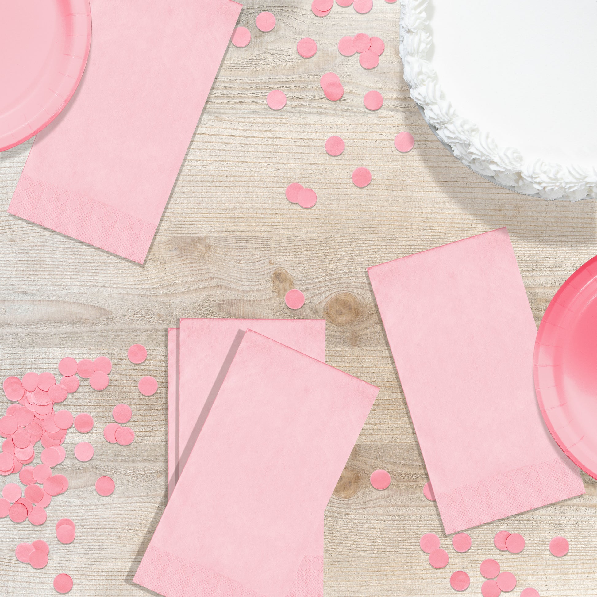 Pink party supplies including napkins and plates on a wooden surface with pink confetti.